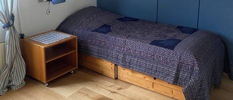 A bedroom with a sloped ceiling, a bed with a patterned bedspread, a small wooden nightstand, a blue lamp, and a poster on the wall.