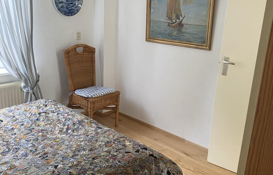 A room with a bed covered in a patterned blanket, a wicker chair, a painting of a sailboat, and a door.