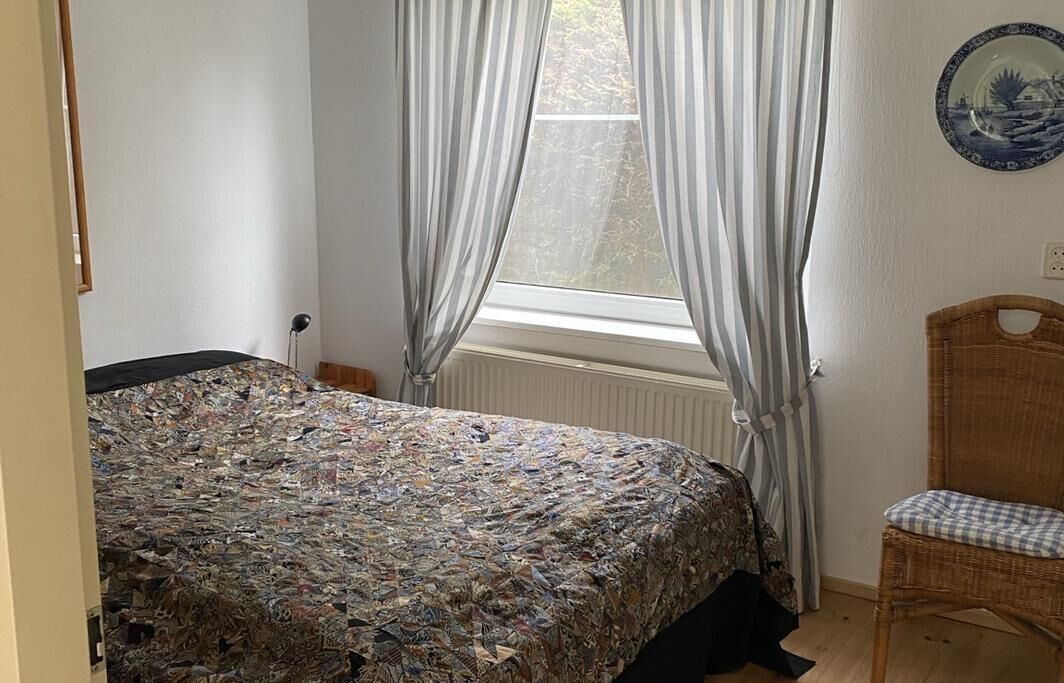 Un dormitorio con cama, una silla, un ventanal con cortinas y un plato fijado a la pared.