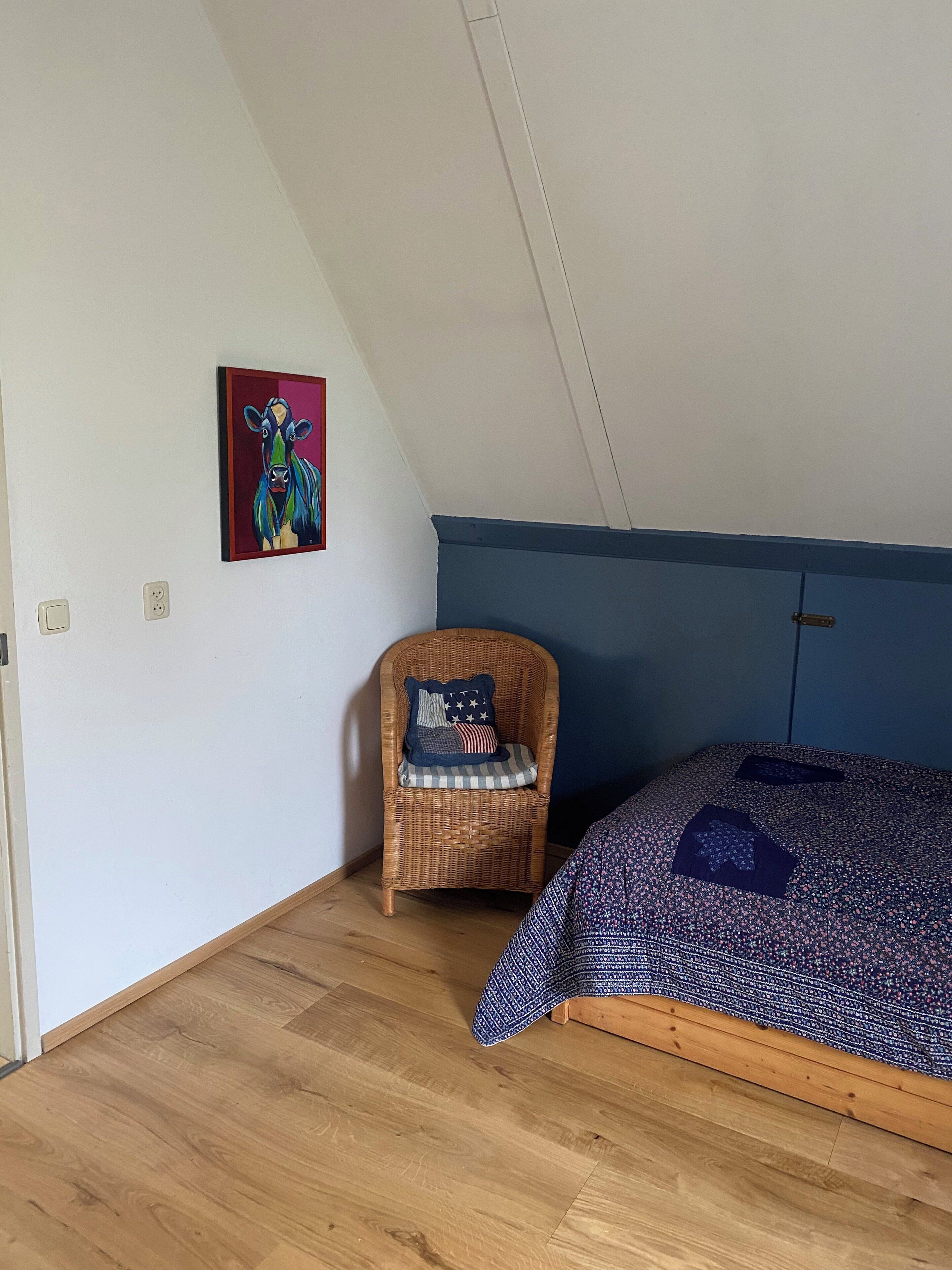 Una habitación con techo inclinado, una cama con colcha estampada, una silla de mimbre con cojín, y un cuadro de colores en la pared.
