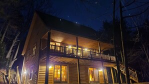A two-story log cabin with a balcony, illuminated by warm interior lighting.