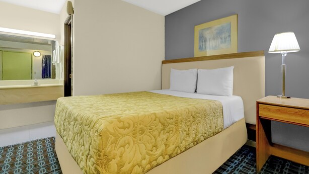 A hotel room with a bed, a bedside table with a lamp, a mirror, and a painting on the wall. - Brookville Inn By OYO Near Dayton International Airport (Brookville)