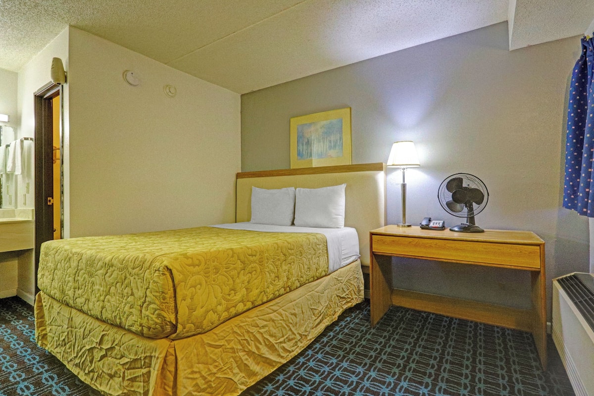 Ein Hotelzimmer mit Bett, Nachttisch, Lampe, Ventilator und einem Bild an der Wand.