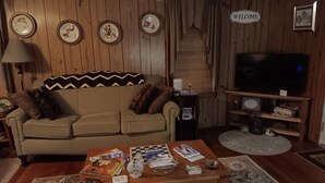 Un salon chaleureux avec un canapé, une table basse et un téléviseur. Des assiettes décoratives ornent le mur, et un panneau d’accueil est visible.