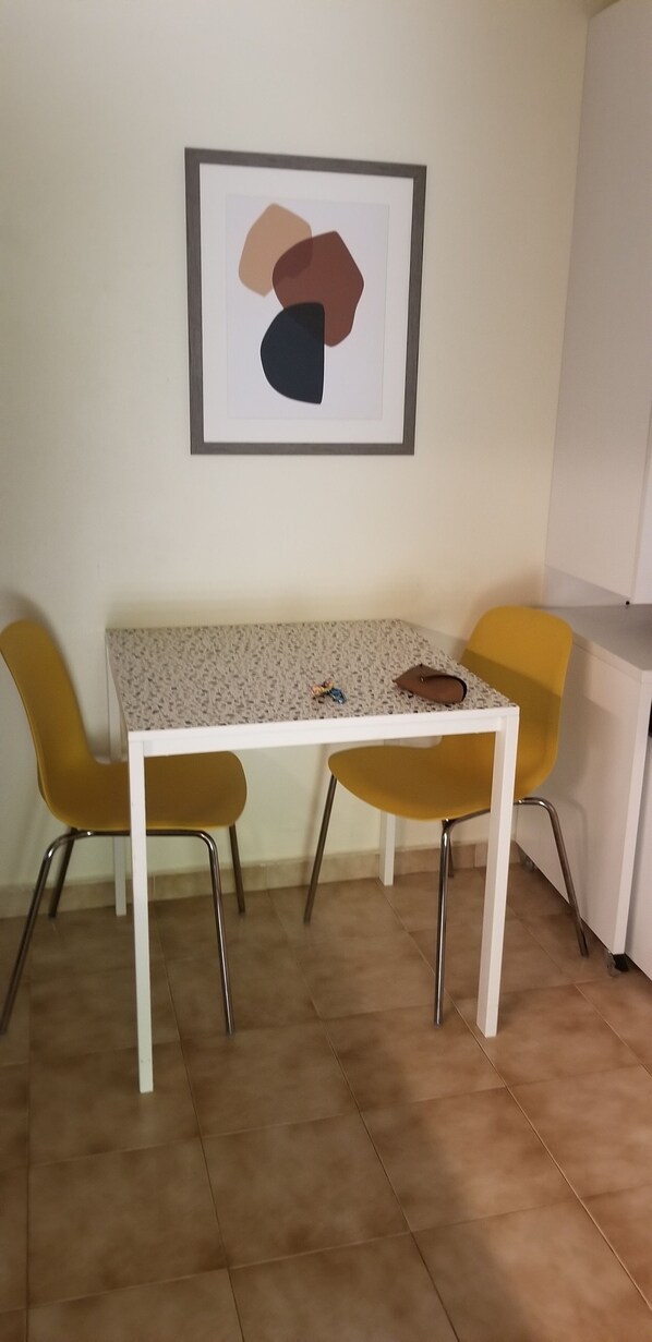 Un comedor con una mesa blanca y dos sillas amarillas, una obra de arte abstracta enmarcada en la pared, y una cocina al fondo.