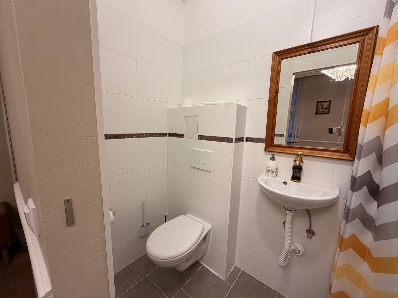Un baño pequeño y limpio con azulejos blancos, inodoro blanco, lavamanos con espejo y cortina de ducha.