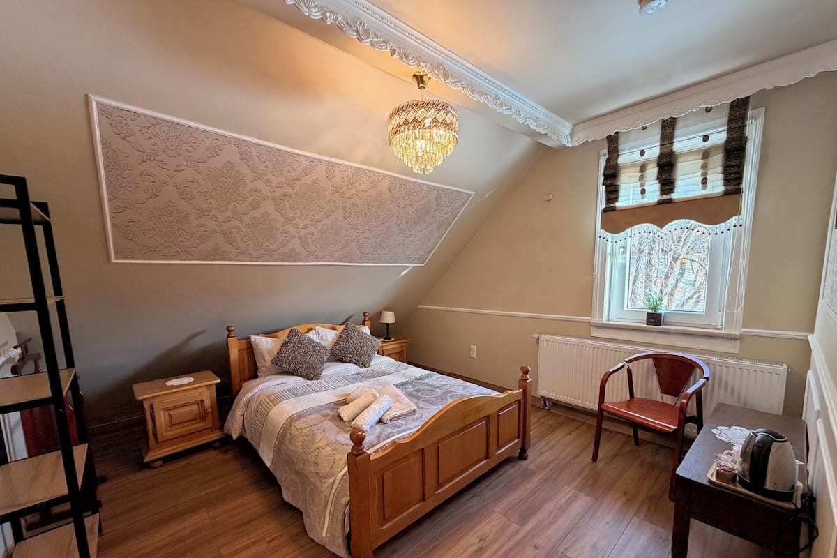 A bedroom with a wooden bed, a chandelier, a radiator, a window with curtains, and a chair.