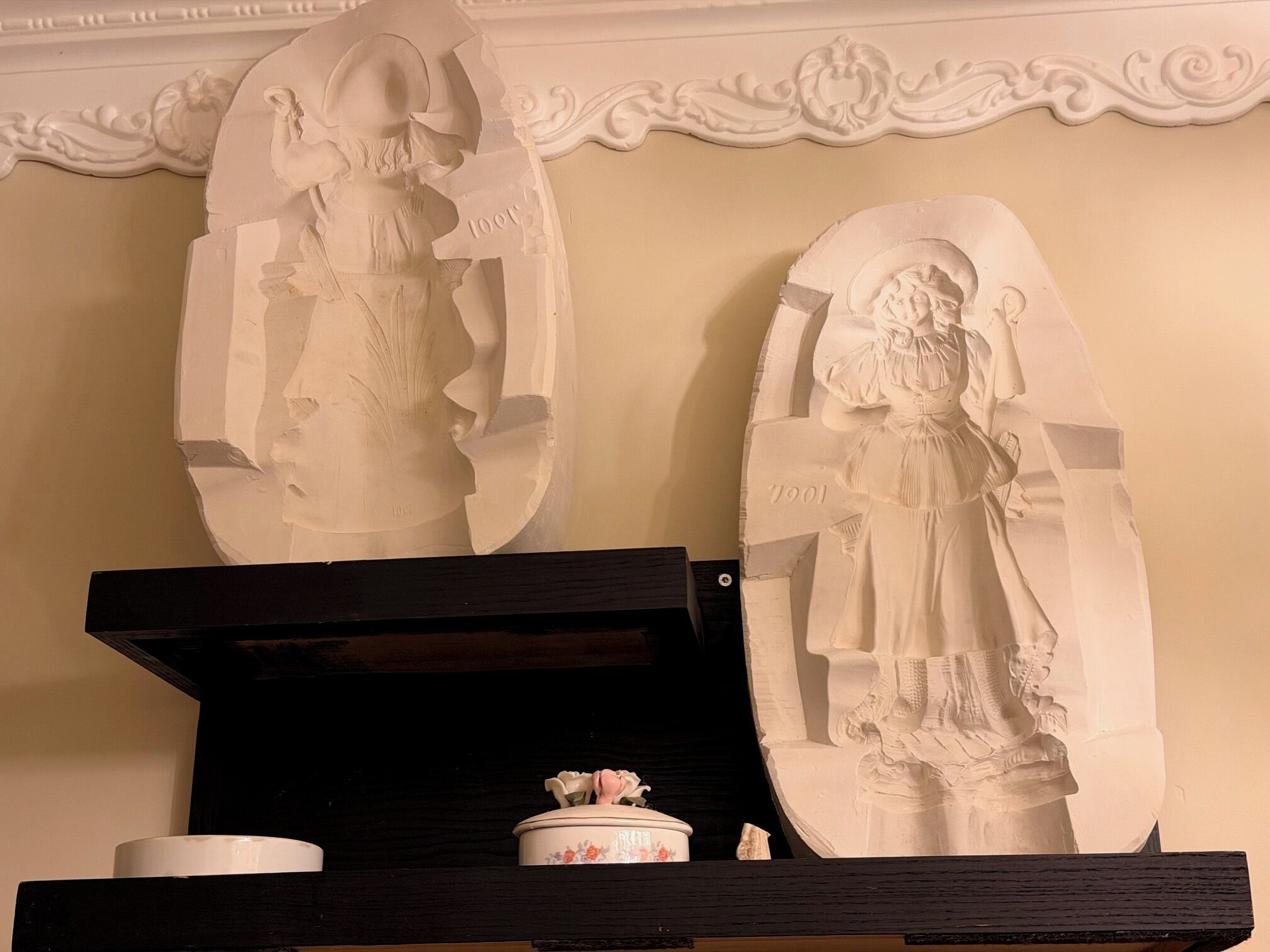 Zwei große, weiße Skulpturen auf einem schwarzen Regal vor einer Wand mit aufwendiger Stuckleiste.