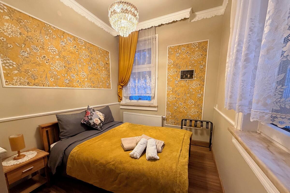 A bedroom with a bed, a nightstand, a chair, a window with curtains, and a chandelier.