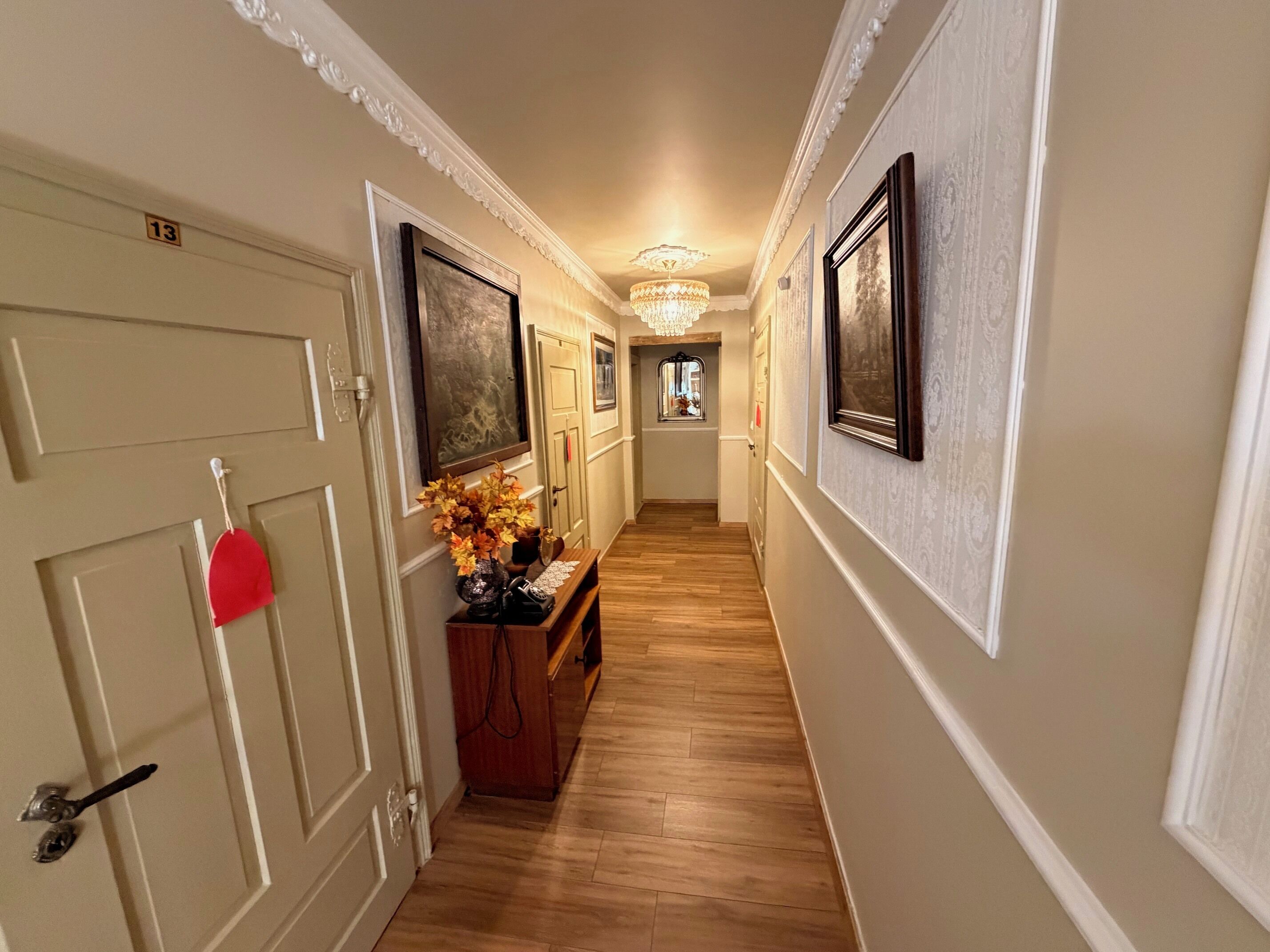 A hallway with wooden flooring, white walls, and framed artwork on the walls. There is a door with a number sign, a bench with a vase of flowers, and a chandelier on the ceiling.