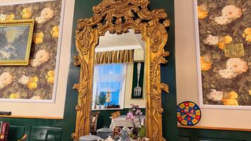 A room with a large ornate mirror, floral wallpaper, a framed painting, a dartboard, and various decorative items.