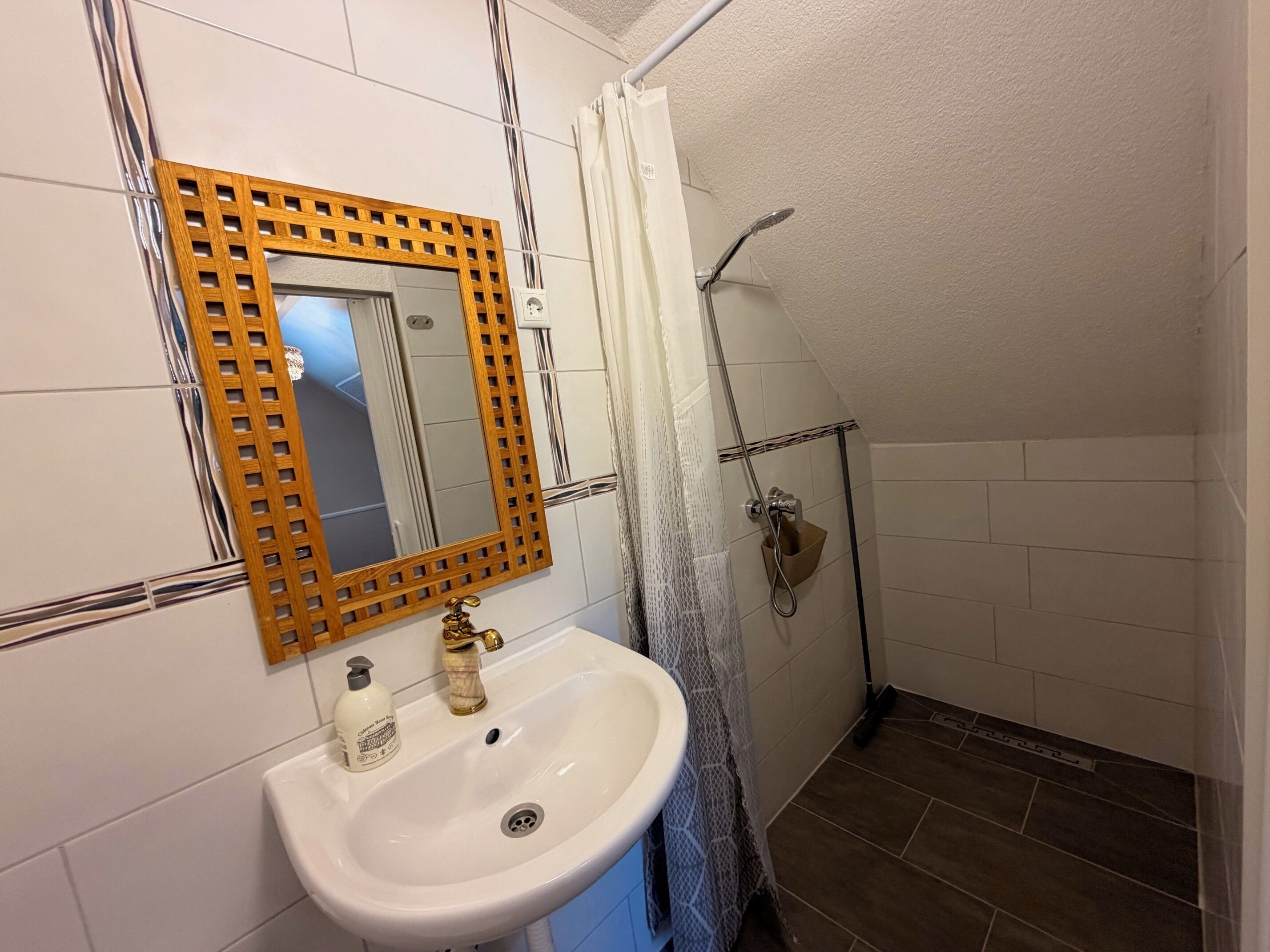 Un baño con lavamanos blanco, espejo con marco de madera y zona de ducha.