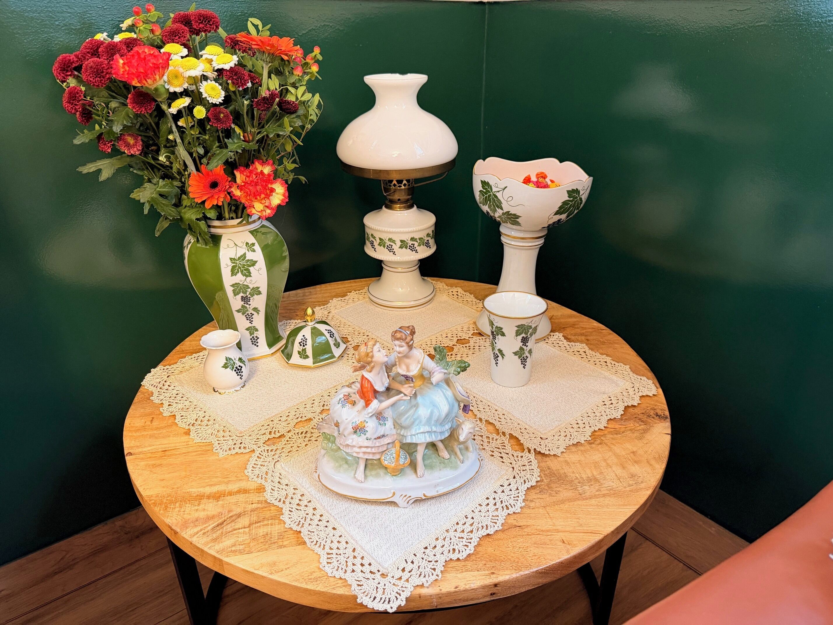 Una mesa redonda de madera con un mantel de encaje, un florero con flores, una lámpara, una pieza decorativa de cerámica y un pequeño cuenco.