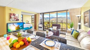 A hotel room with a dining area, a sofa, a TV, and a balcony with a city and mountain view. - Waikiki Banyan 1BR Condos with No Parking (Honolulu)