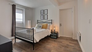 Un dormitorio con estructura de cama metálica negra, ropa de cama blanca, una mesita de noche con lámpara y una ventana con persianas.
