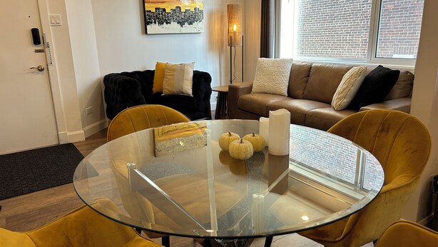 Una sala de estar moderna con una mesa de vidrio, sillas amarillas, un sofá marrón y un cuadro de un paisaje urbano en la pared.