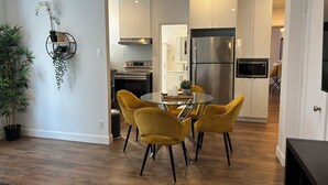 Una cocina moderna con armarios blancos, un refrigerador de acero inoxidable y un microondas. Hay un comedor con una mesa de vidrio y sillas amarillas.