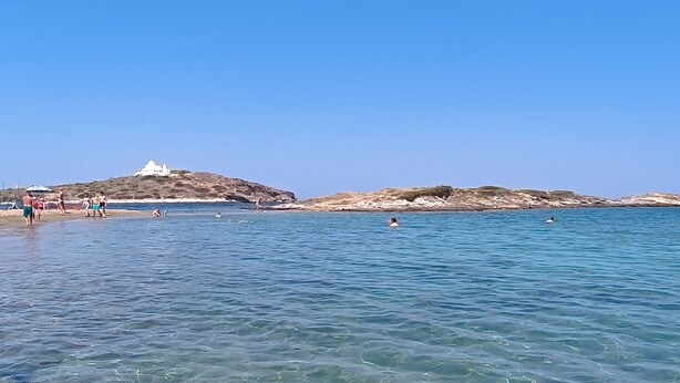 Una zona costera con aguas poco profundas y cristalinas, una playa de arena y terreno rocoso al fondo.