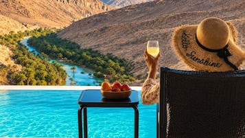 En person som kopplar av vid en pool med utsikt över en flod och berg, och som håller i ett glas och en skål med frukt.