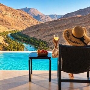 A person relaxing by a pool with a view of a river and mountains, holding a glass and a bowl of fruit. - WADINA RESORT   (Wadi Bani Khalid)