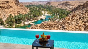 A pool with a table in the foreground, a view of a river, and rocky terrain in the background. - WADINA RESORT   (Wadi Bani Khalid)