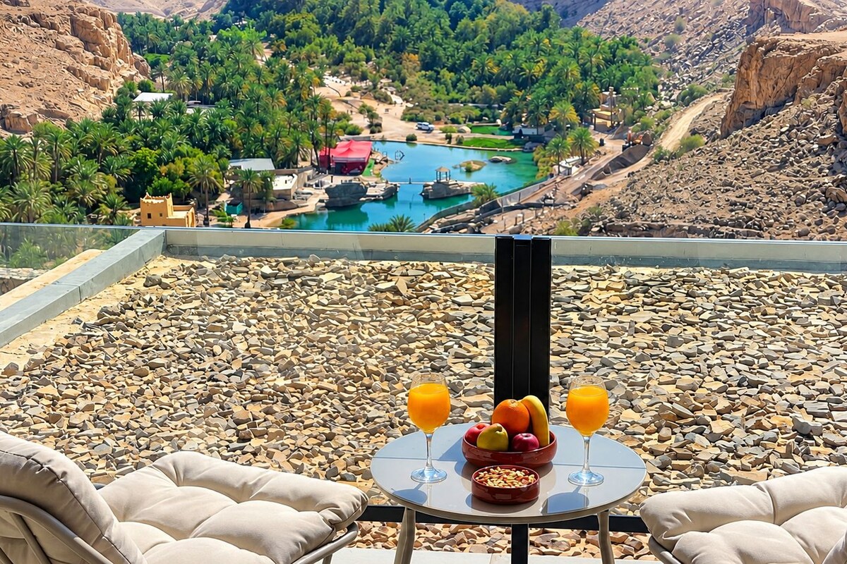 A terrace with a table set for breakfast, overlooking a mountainous landscape with a pool and buildings.