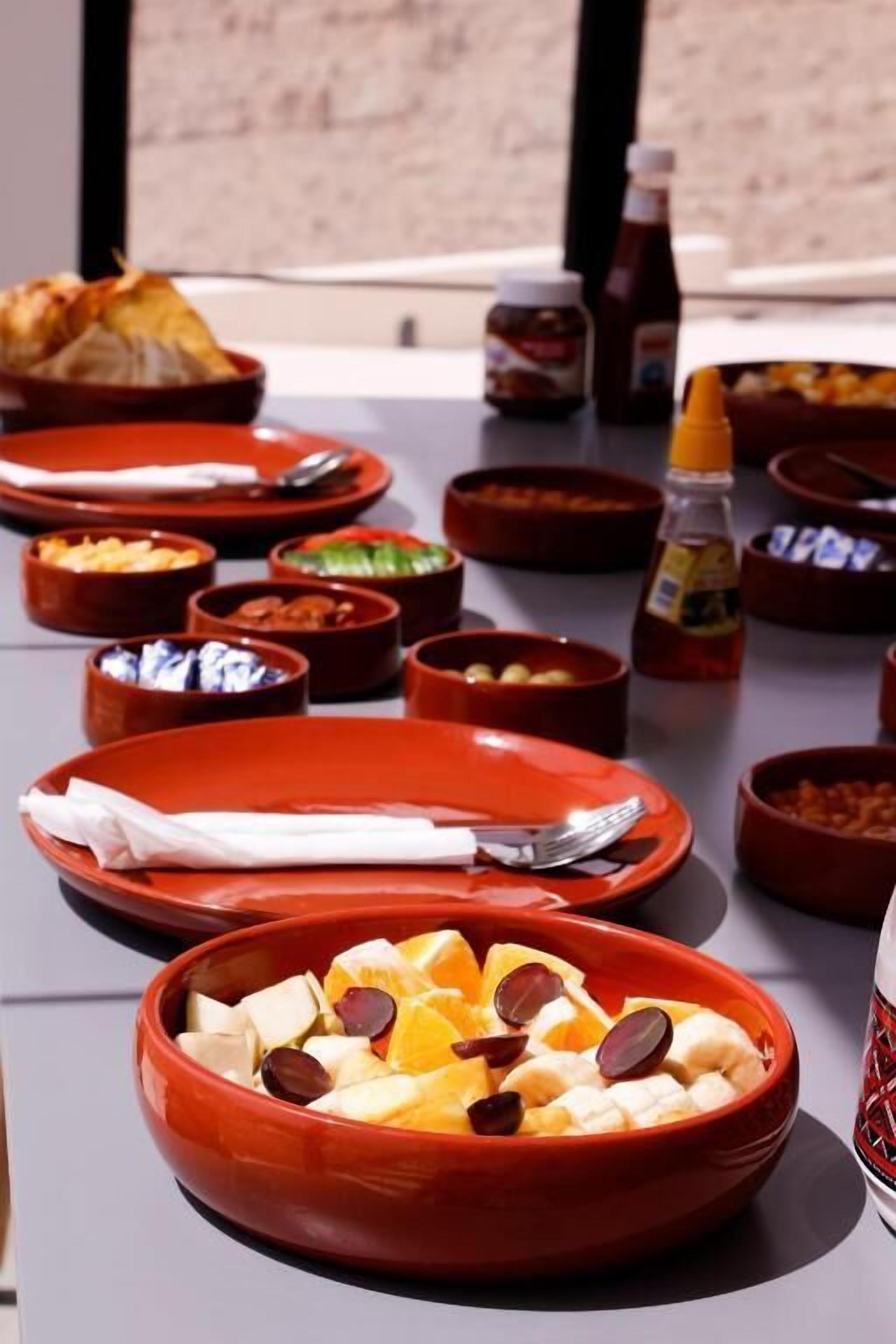 A dining table with multiple red bowls containing various foods, a bottle of sauce, and a basket of bread.