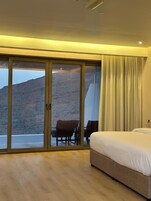 Una habitación de hotel con un amplio ventanal corredero que da a un balcón, una cama y una zona de estar con una mesa y sillas.