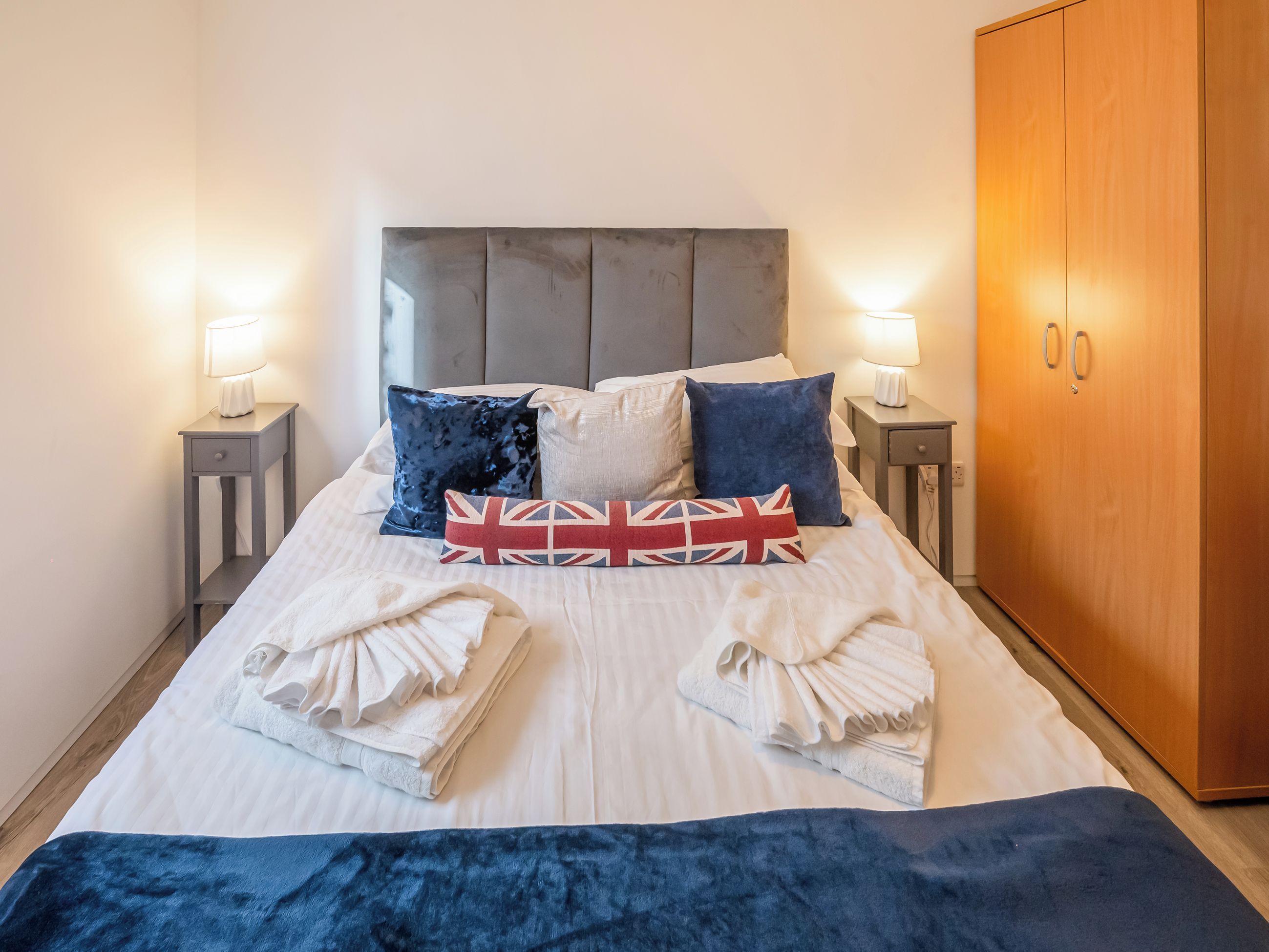 A neatly made bed with pillows, a headboard, bedside tables with lamps, and a wardrobe.