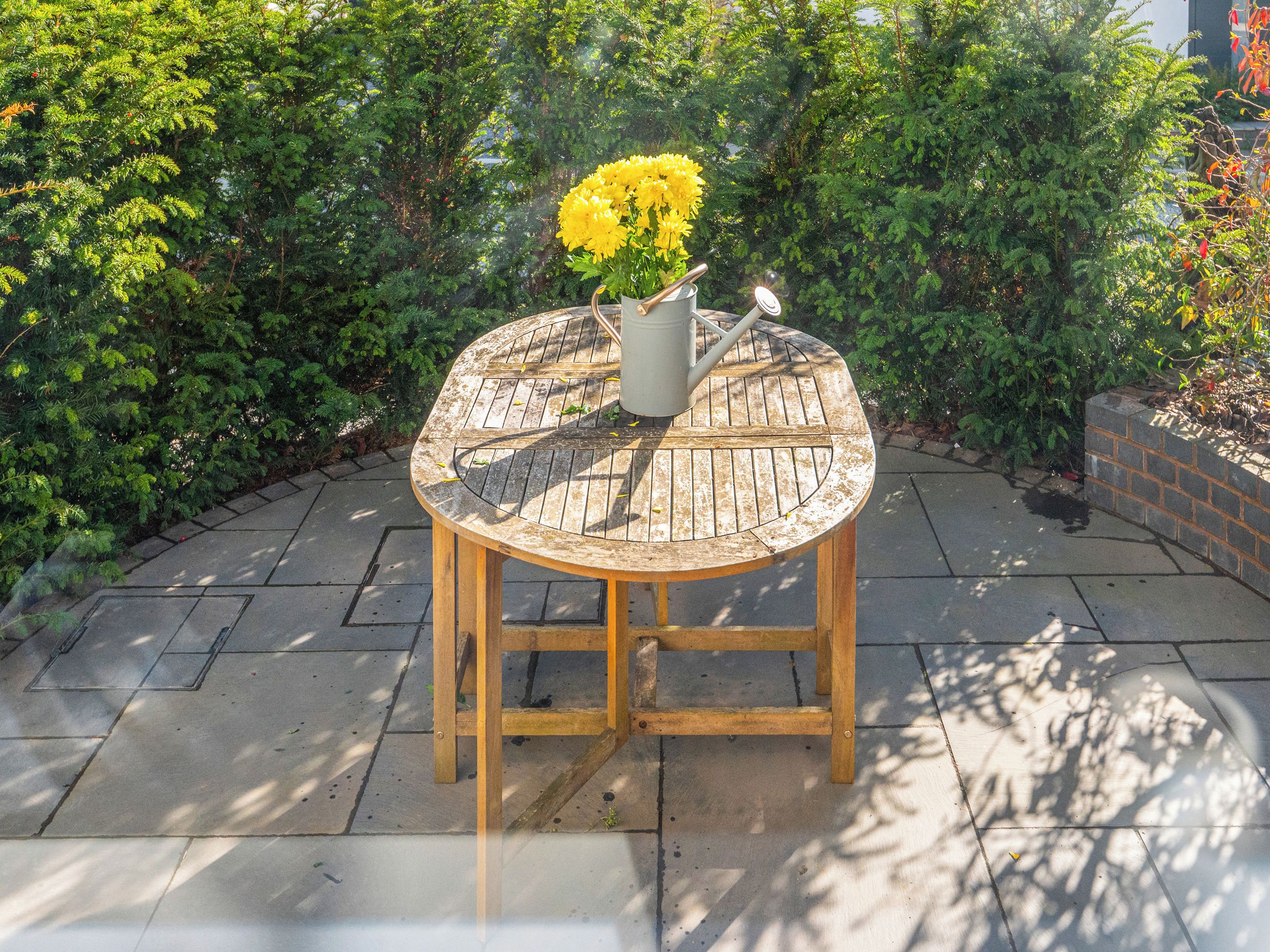 Une table ronde en bois, sur laquelle sont posés un pichet en métal et des fleurs, le tout entouré de verdure.