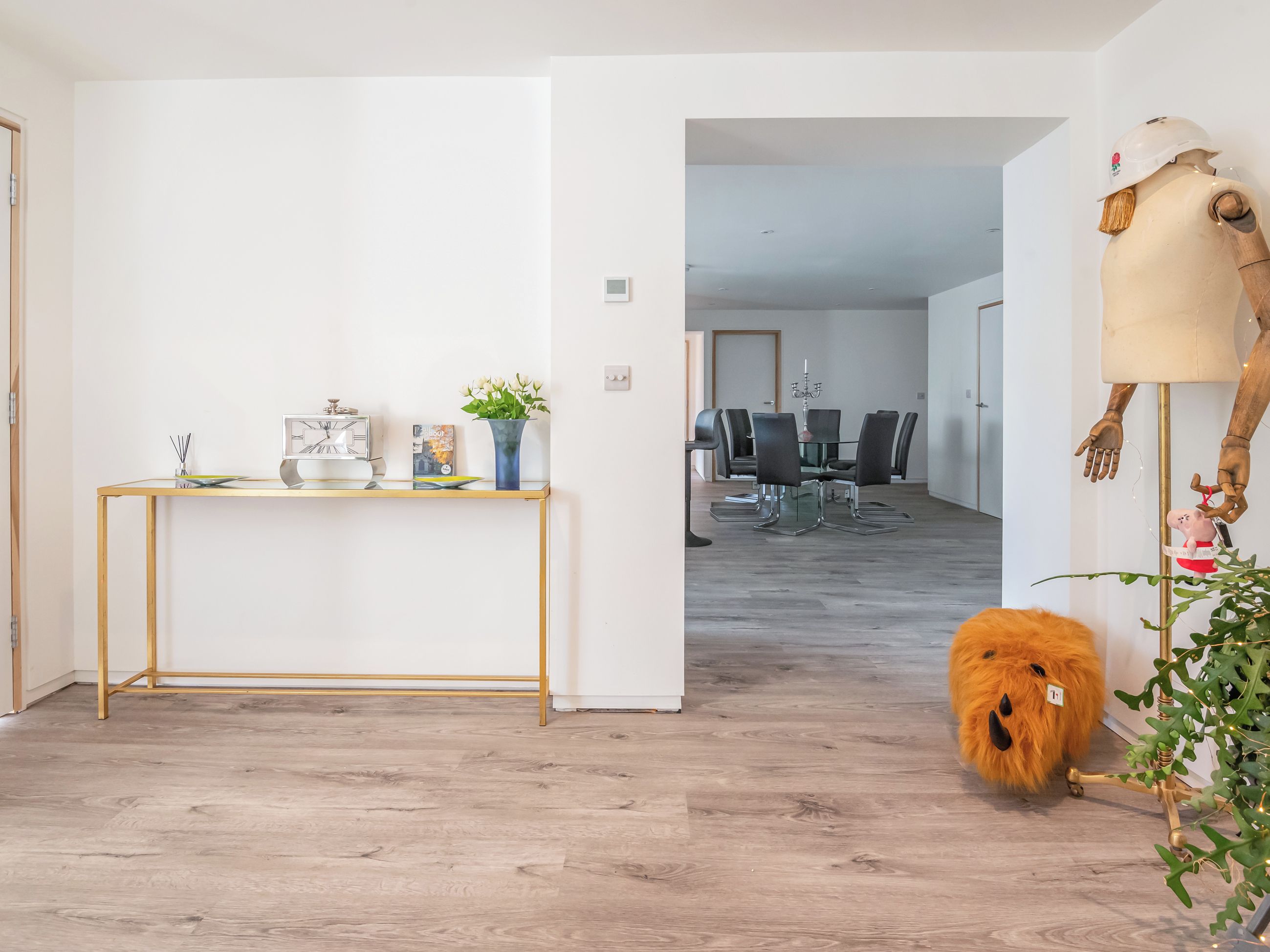A modern interior with a wooden floor, a glass-top console table, a dining area with a chandelier, and a unique wooden sculpture.
