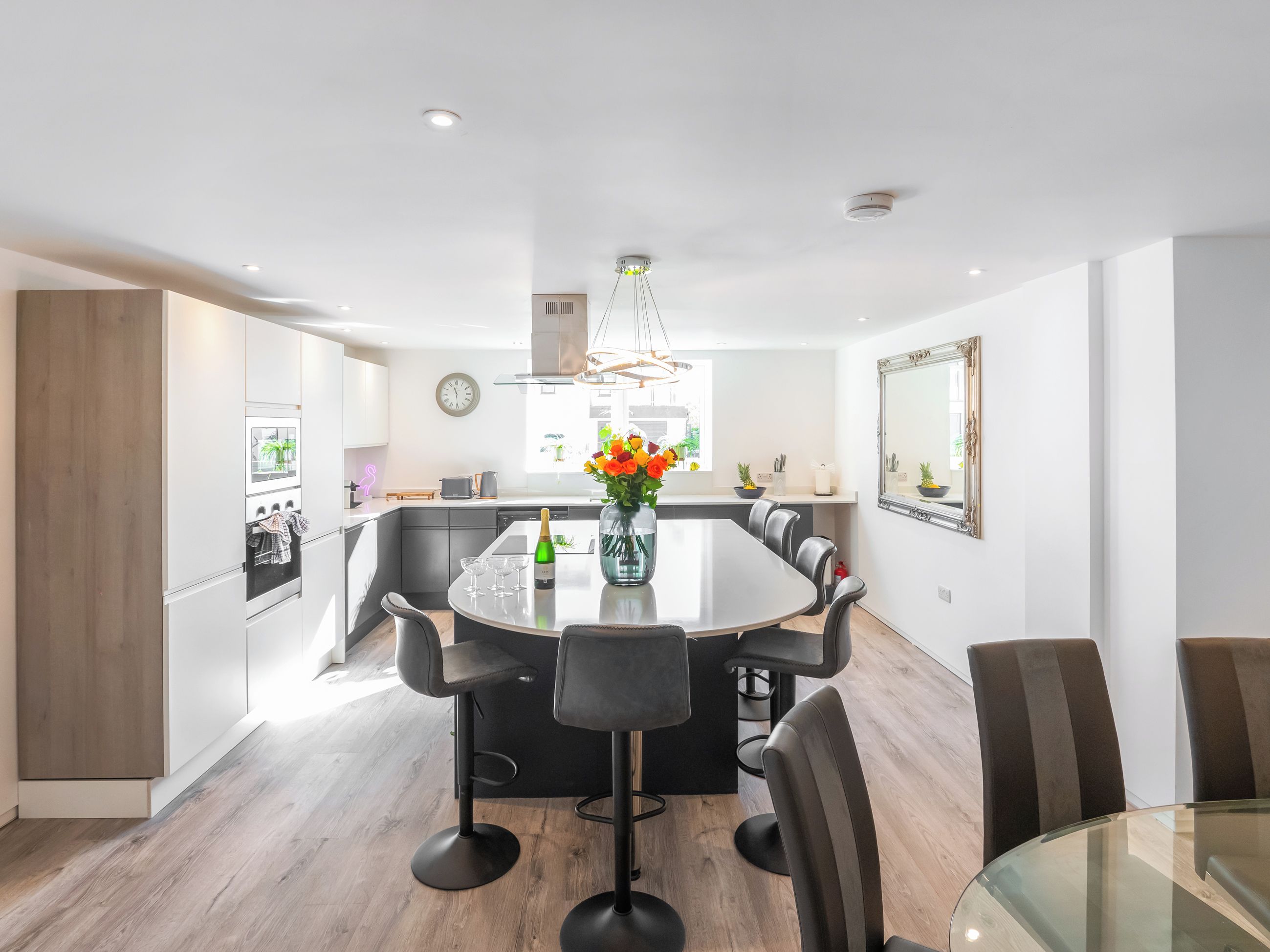 A modern kitchen with a central island, black bar stools, a dining table with chairs, and a large mirror on the wall.