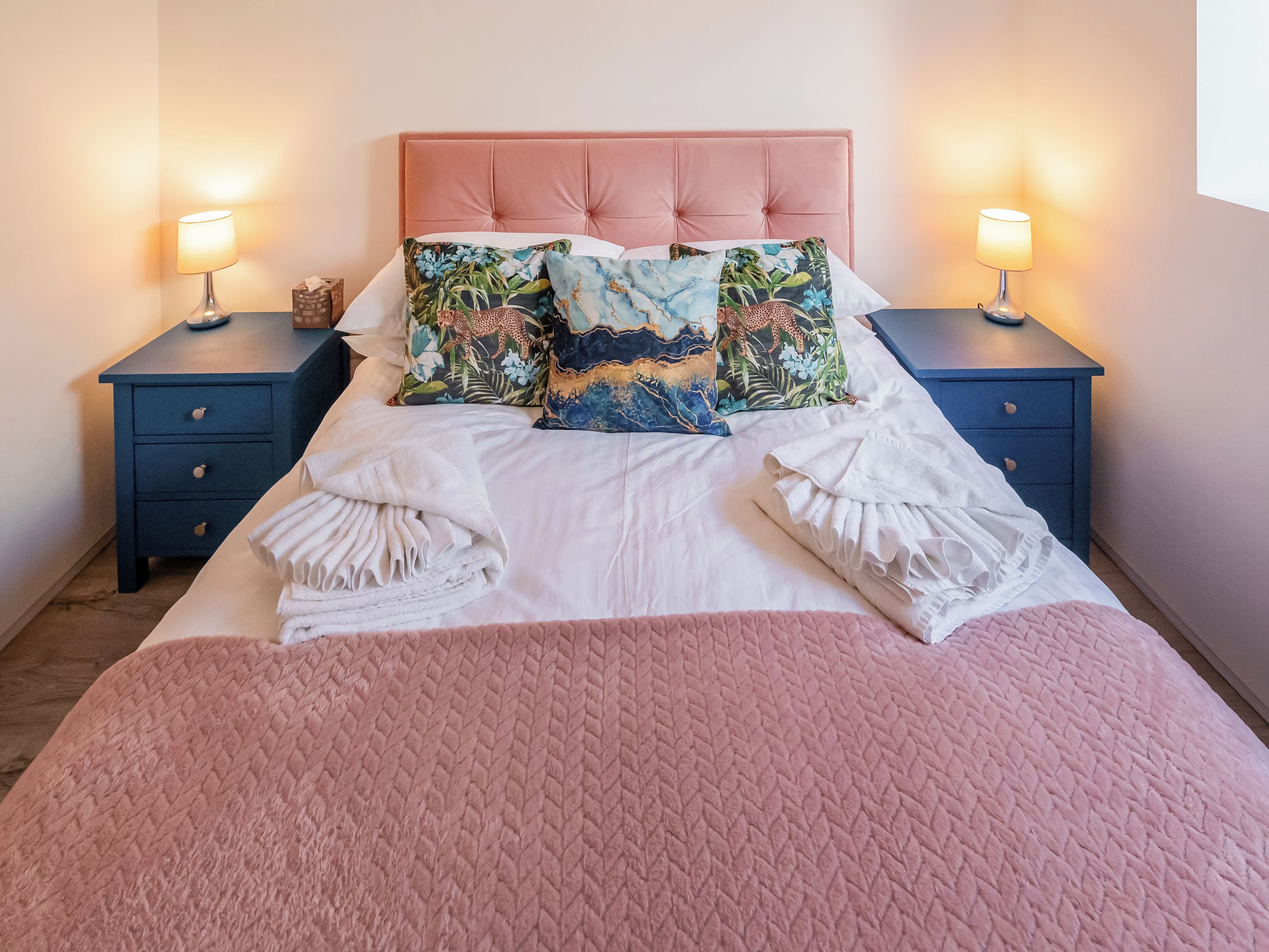Una cama bien hecha con colcha rosa, dos mesitas de noche con lámparas y almohadones decorativos.