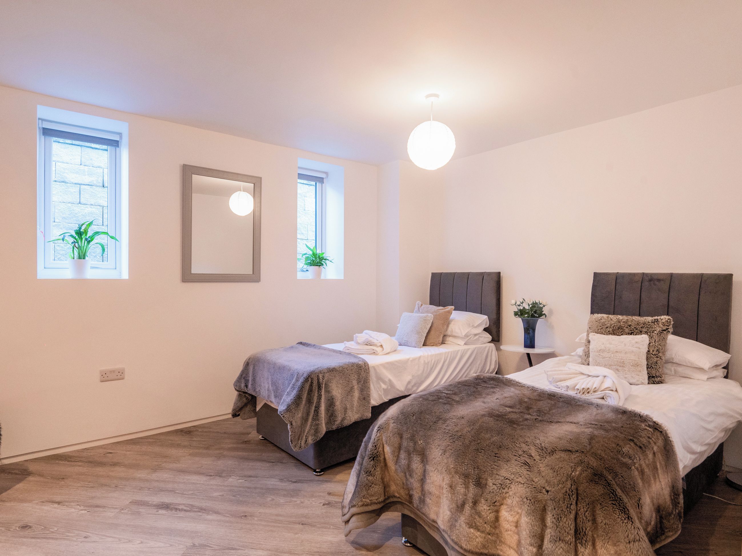 A bedroom with two beds, a bedside table with a lamp, a window with a plant, and a mirror on the wall.
