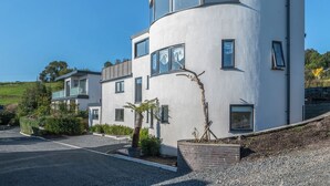 Une maison moderne à deux étages, dotée d’une façade incurvée, de grandes fenêtres et d’un petit jardin agrémenté d’un arbre et d’arbustes.