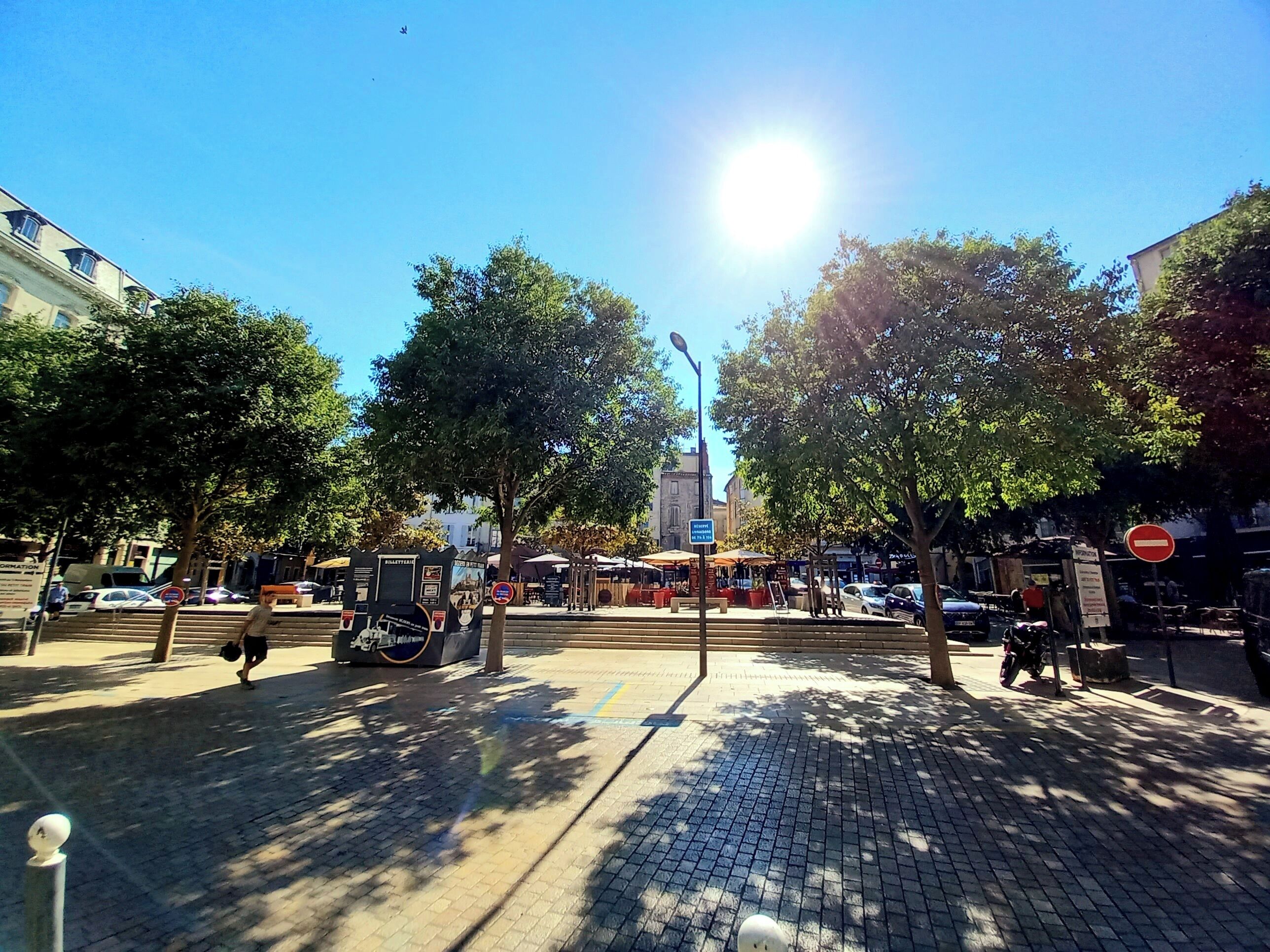 Eine sonnige Stadtstraße mit Bäumen, einem Verbotsschild und einem Gebäude mit einem Glockenturm im Hintergrund.