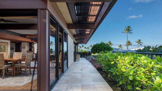 A modern outdoor dining area with a view of the ocean, featuring a wooden table and chairs, a glass-enclosed patio, and tropical landscaping. - Ocean View Villa w/ Close Proximity to Ocean and Resort Amenities (Waimea)