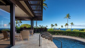 A modern outdoor pool area with a view of the ocean, furnished with wicker chairs and a glass-enclosed patio. - Ocean View Villa w/ Close Proximity to Ocean and Resort Amenities (Waimea)