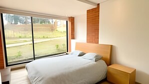 Ein modernes Schlafzimmer mit einem großen Bett, einem Holzkopfende und Blick auf eine Rasenfläche durch eine Schiebeglastür.