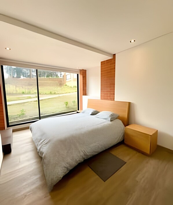 Une chambre moderne avec un grand lit, une tête de lit en bois et une vue sur un espace verdoyant, visible à travers une porte-fenêtre coulissante.
