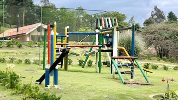 A playground with various climbing and sliding structures, surrounded by a grassy area and trees.