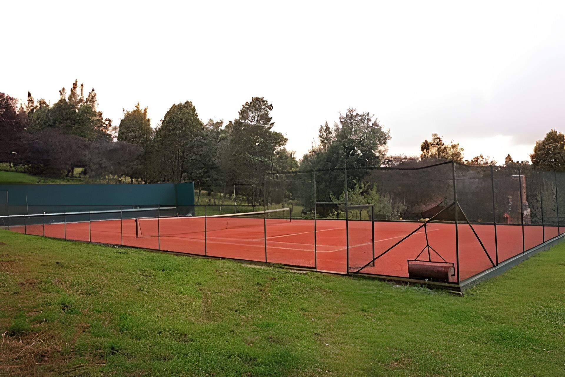Ulkotenniskenttä, jossa on vihreä aita, ja jota ympäröi nurmikko ja puita.