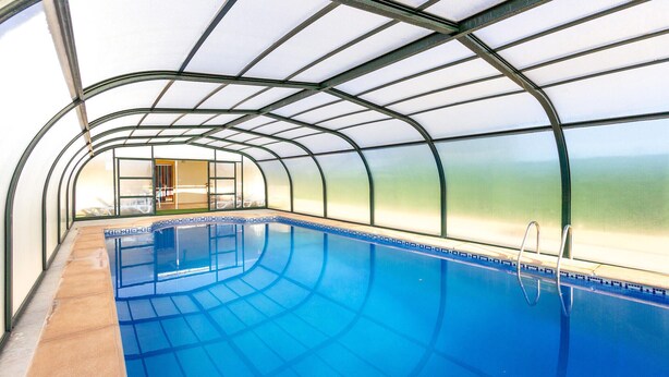 An indoor swimming pool with a glass ceiling, clear blue water, and a pool ladder. - Casa Rural el Olivo centenario (Perin)