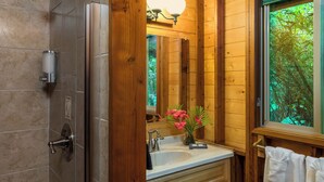 A bathroom with a wooden interior, a white sink, a mirror, and a window with a view of greenery. - Private Rainforest Retreat in Volcano Village (Volcano)