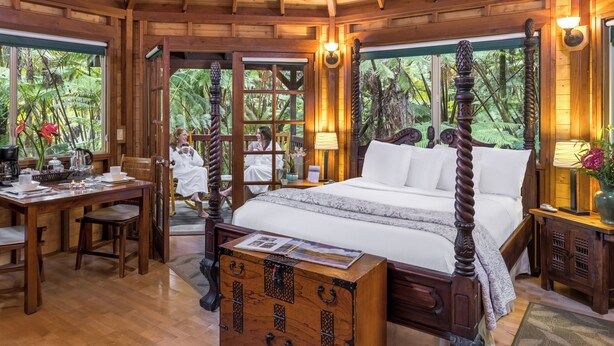 A wooden four-poster bed with a wooden chest of drawers in front, a dining table with chairs, and a view of lush greenery through large windows. - Rainforest Retreat in Volcano Village (Volcano)