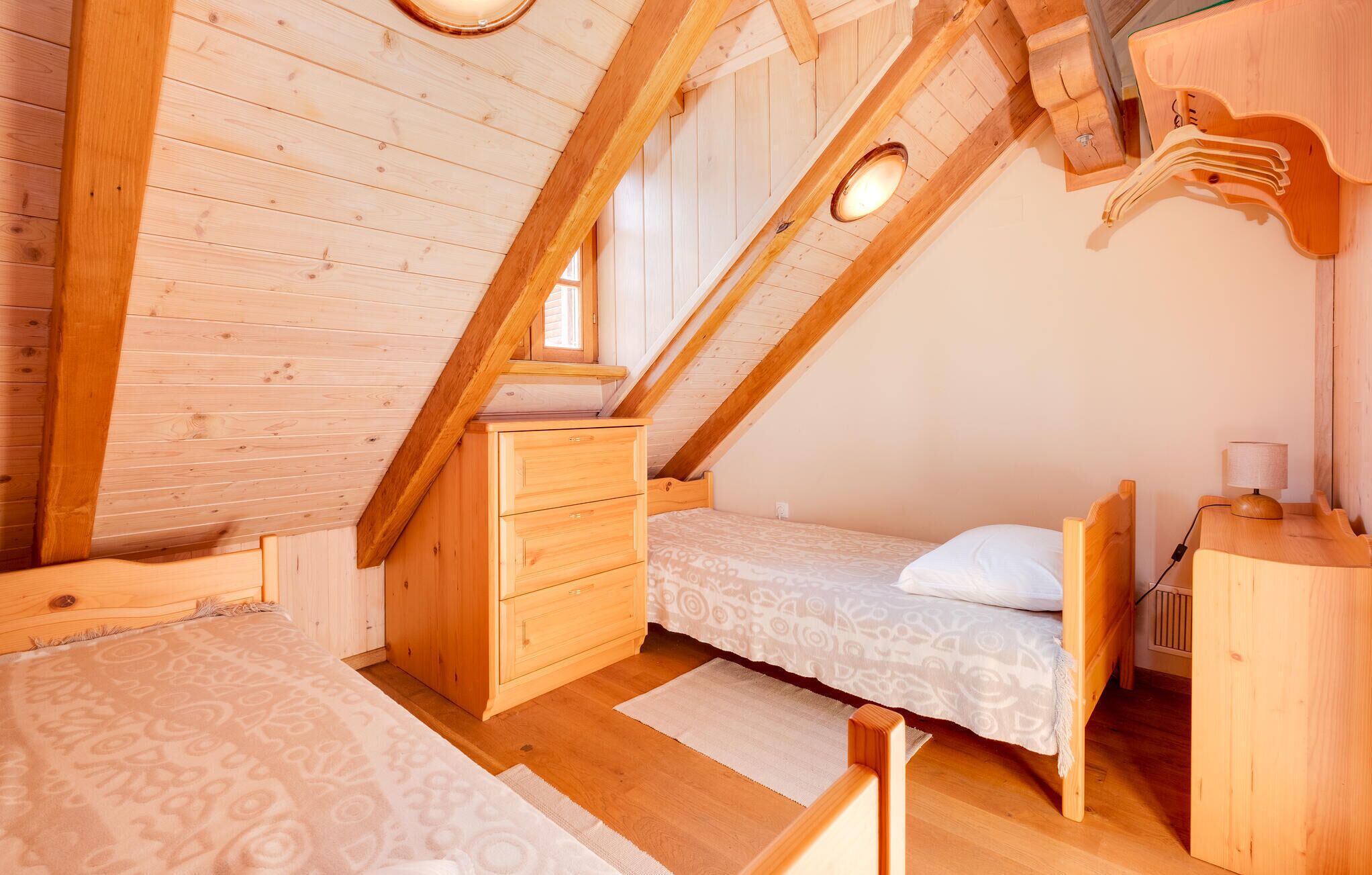 A cozy attic bedroom with wooden walls and a sloped ceiling. It includes a bed, a dresser, and a small desk with a lamp.