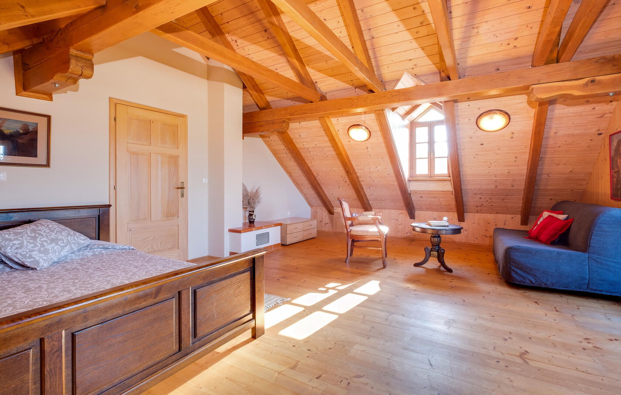 A cozy attic room with a wooden ceiling, a bed, a blue sofa, and a small table.