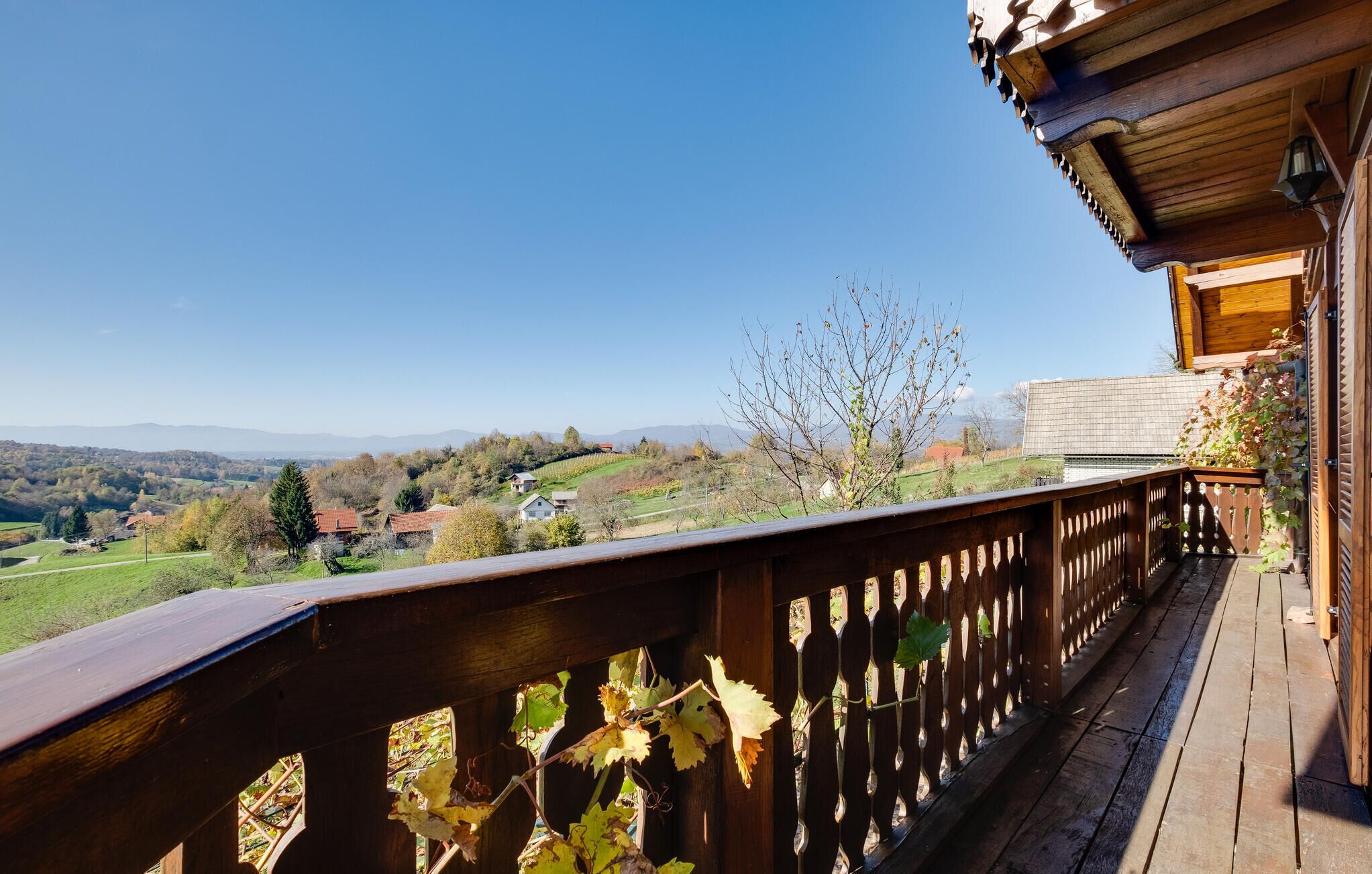 A wooden balcony with a view of a rural landscape, including houses and mountains in the distance.