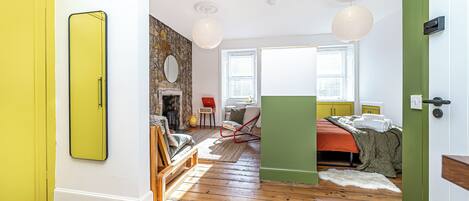 A room with a wooden floor, a bed with a green bedspread, a red chair, a small table with a vase, and a mirror on the wall.