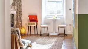 A cozy room with a wooden floor, a white wall, a window with blinds, a small round table with two stools, a red box, and a yellow lamp. - Funky Studio Near The Beach (Burntisland)